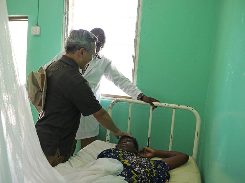 Durante una visita a la clínica y dispensario de Ndoleleji que asiste a unos 40 pacientes por semana, el padre Dinh, acompañado por un oficial clínico, bendice a una mujer enferma. (Sean Sprague/Tanzania)