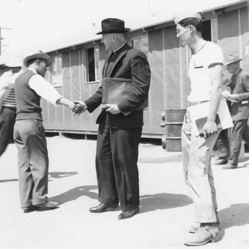 japoneses-estadounidenses:El padre Lavery le da un apretón de manos a un interno en el campamento Manzanar. (Maryknoll Mission Archives)