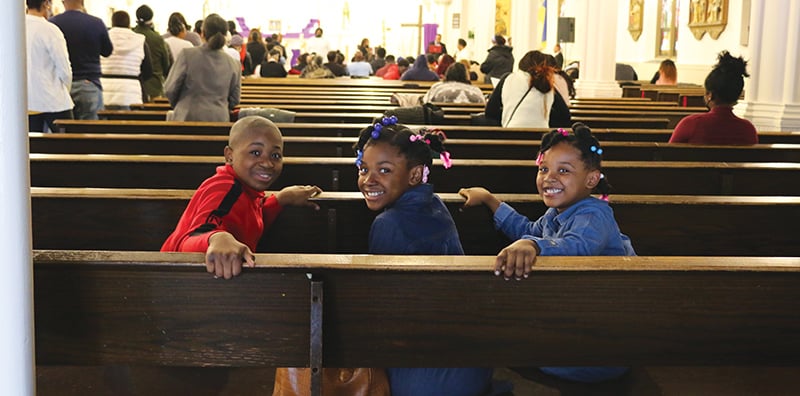 Los niños Anderson, Anchina y Briana Brice sonríen sentados en una banca de la iglesia mientras sus padres, Andre e Ilna D. Brice, van a recibir la Comunión en la celebración mensual. (Diane Mastrogiulio/EE.UU.)